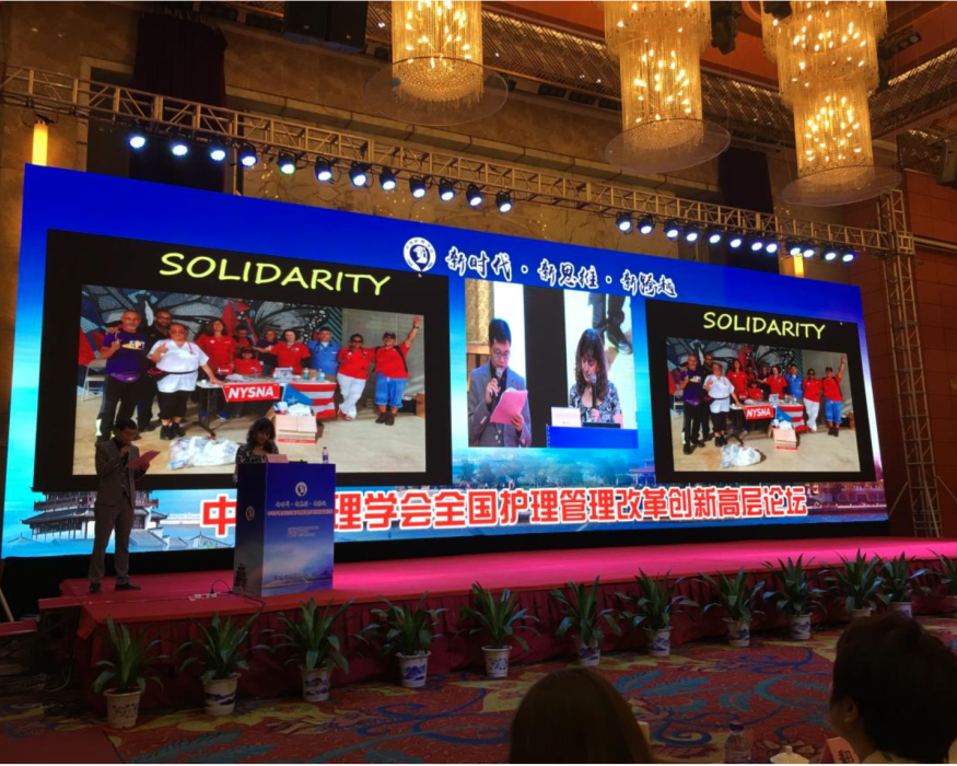 Large Monitor at a Convention shows Judy Sheridan Gonzalez speaking to the crowd at Beijing